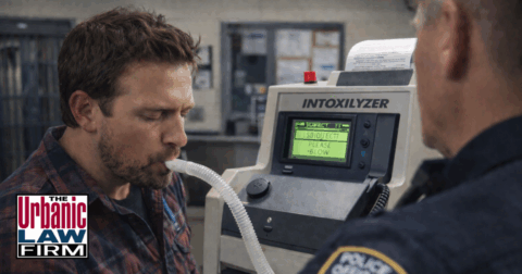 Daytime photo-style image of a man in an Oklahoma police station blowing into an Intoxilyzer machine while an officer looks on, illustrating high-stakes DUI arrests and related obscene or threatening communication criminal defense handled by The Urbanic Law Firm.