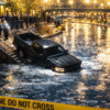 Nighttime photo-style image of a dark pickup truck nose-down in the icy Bricktown canal in Oklahoma City, brightly lit by streetlamps, with “police line do not cross” tape, officers on scene, and an Oklahoma criminal defense attorney from The Urbanic Law Firm talking with a worried driver about DUI charges.