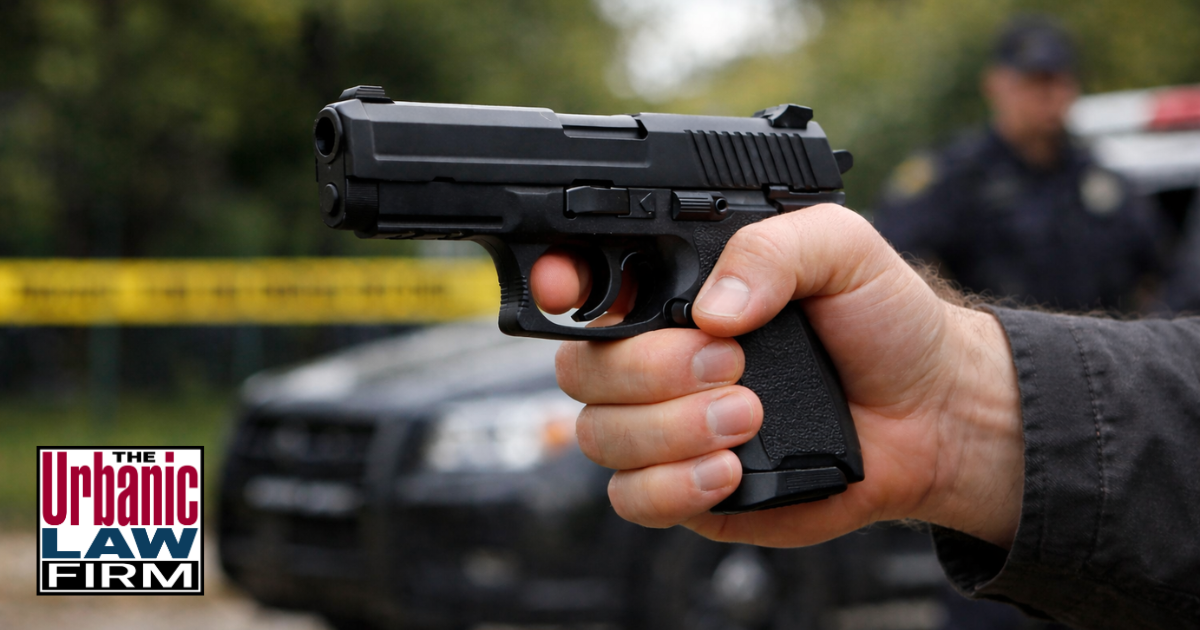 Daytime close-up photograph of an adult hand gripping a handgun inside a vehicle on an Oklahoma street, symbolizing supplying firearms to prohibited persons in Oklahoma and other serious gun charges, with the scene illustrating the need for experienced Oklahoma criminal defense representation by The Urbanic Law Firm.