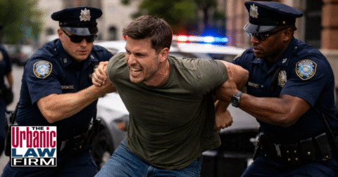 Daytime photo-style image of a tense Oklahoma street scene where a young man appears to be resisting arrest as uniformed police officers attempt to detain him beside a patrol SUV, symbolizing an Oklahoma resisting arrest criminal defense attorney protecting the accused and illustrating Oklahoma criminal defense representation by The Urbanic Law Firm.