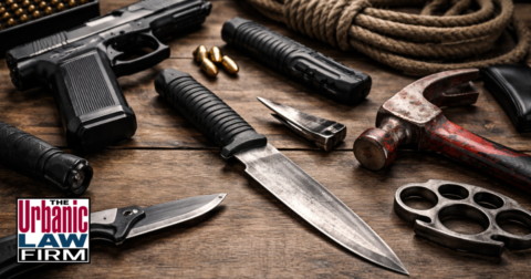 Deadly weapon assault Oklahoma image showing a handgun, knives, hammer, brass knuckles, rope, and ammunition on a wooden surface, illustrating Oklahoma criminal defense by The Urbanic Law Firm.